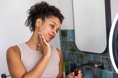 African american woman applying facial serum at bathroom vanity holding amber dropper bottle