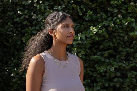 Sunlit reflective woman standing by lush hedge wearing sleeveless ribbed top and gold chains
