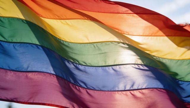 Waving rainbow pride flag glowing in bright sunlight against clear blue sky