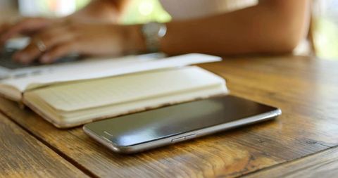 Smartphone resting on wooden desk with notebook and laptop, sunlit home workspace