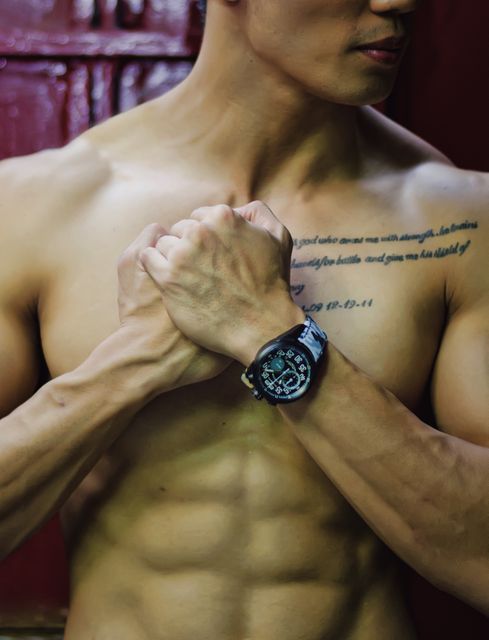 Muscular man without a shirt clasping hands in front of muscular torso, showing wristwatch and tattoo. Ideal for fitness, bodybuilding, and men's health advertisements, fitness blogs, motivational posters and sports-related content. Emphasizes strength, masculinity, and physical fitness.