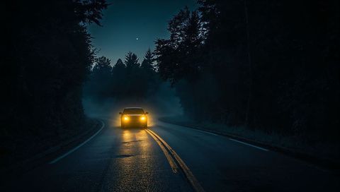 Car cutting through fog on dark forest road with reflective double yellow lines