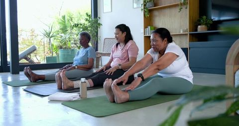Mature diverse women stretching on yoga mats together in bright living room with plants