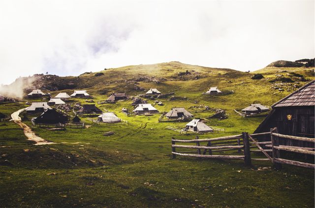Traditional wooden houses surrounded by lush green hills in an idyllic Alpine village. Gentle mist adds a serene touch while the inviting scene showcases untouched nature. Perfect for travel brochures, nature blogs, and illustrations promoting eco-tourism or rural retreats.
