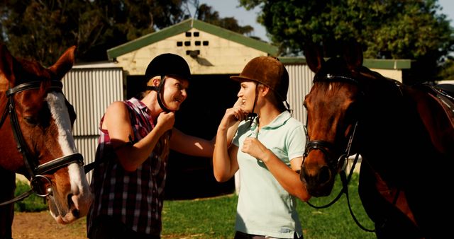 Two equestrians, in casual attire and riding helmets, engage in a cheerful conversation while preparing their horses for a ride. The setting is a rustic outdoor barn, ideal for themes of friendship, outdoor activities, horse training, and teamwork. Perfect for use in ads, articles, and content related to equestrian sports and hobbies, outdoor adventures, or rural life.