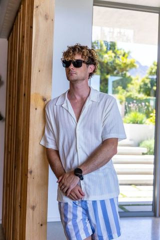 Curly-haired man leaning on wooden column in sunlit modern home wearing sunglasses and striped short