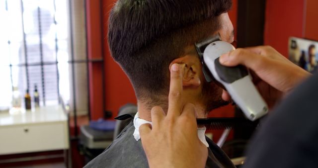 A young Caucasian man is getting a haircut from a barber, with copy space. Precision and care are evident as the barber shapes the man's hairstyle with electric clippers.