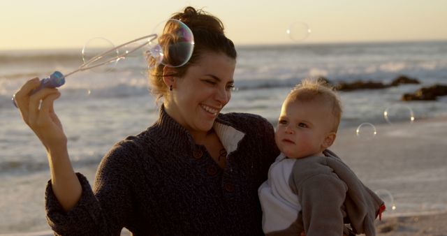 Happy caucasian mother blowing bubbles and carrying toddler son on beach at sunset. Motherhood, childhood, care, free time, travel, nature and healthy outdoor lifestyle, unaltered.
