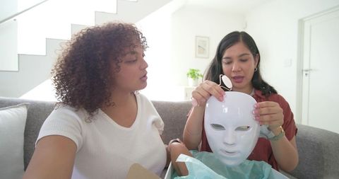 Multicultural friends unboxing led facial mask on couch, exploring home skincare device