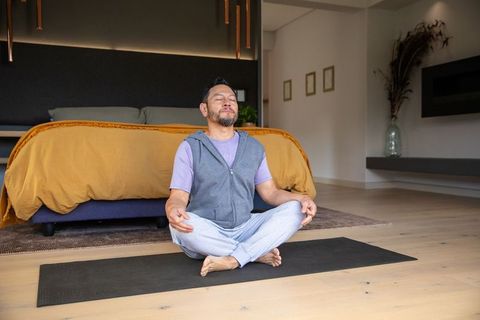 Mature asian man meditating cross-legged on yoga mat in bedroom with mustard duvet