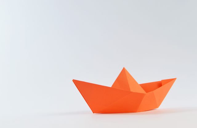 Minimalistic orange paper boat displayed against white background. Perfect for use in educational materials, craft tutorials, creative project backgrounds, inspiration for origami enthusiasts, or promotional content for DIY activities and workshops.