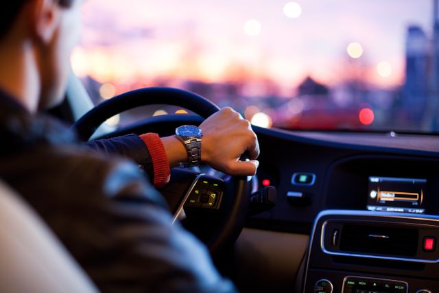 Man wearing smartwatch driving car at sunset with city lights in the background. Car dashboard and steering wheel visible, representing evening commute. Suitable for themes related to transportation, urban lifestyle, or technology in everyday life.
