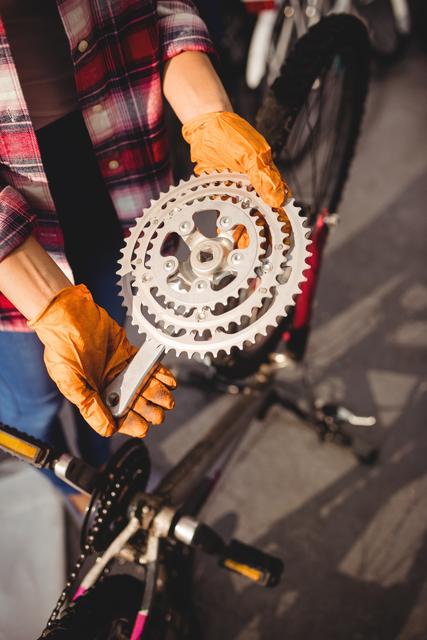 Person wearing gloves holding a bicycle gear in a workshop. Ideal for content related to bicycle maintenance, bike repairs, cycling parts, and mechanical services. Useful for promoting bike shops, mechanic services, and cycling gear maintenance tutorials.
