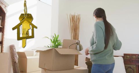 Young woman unpacking boxes in sunlit living room with golden house key charm