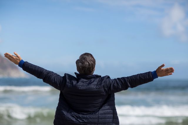Mature man standing on beach with arms outstretched, facing ocean. Ideal for themes of freedom, relaxation, and connection with nature. Suitable for travel brochures, wellness blogs, and inspirational content.