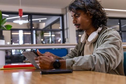 Young Indian man using tablet in modern coworking lounge wearing white headphones and smartwatch
