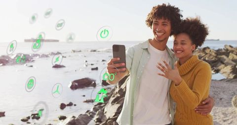 Young couple celebrating engagement on rocky beach taking selfie with ar eco icons
