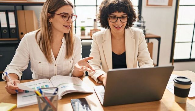 Collaborating women smiling while reviewing charts on laptop and binder at modern office desk