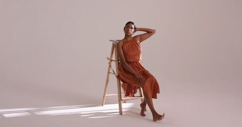 Elegant woman in rust dress sitting on wooden ladder in minimal studio