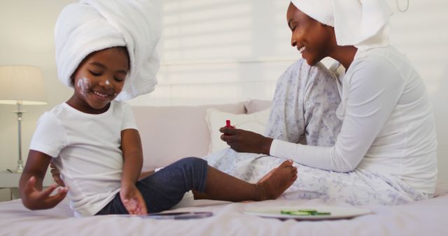 A mother and daughter are spending quality time together, sitting on a bed with face masks and towels wrapped around their heads. The mother is applying nail polish while the daughter looks at her hands, smiling. This image can be used for themes related to family bonding, self-care routines, skincare products, and home life.
