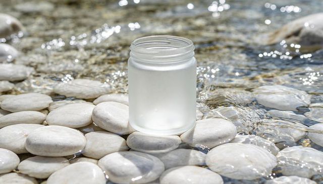 Frosted glass jar sitting on sunlit river pebbles with sparkling reflections and gentle ripples