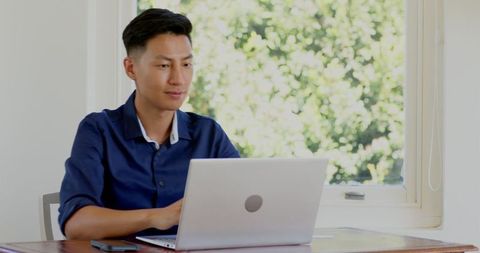 Asian Man Smiling While Using Laptop Near Bright Window