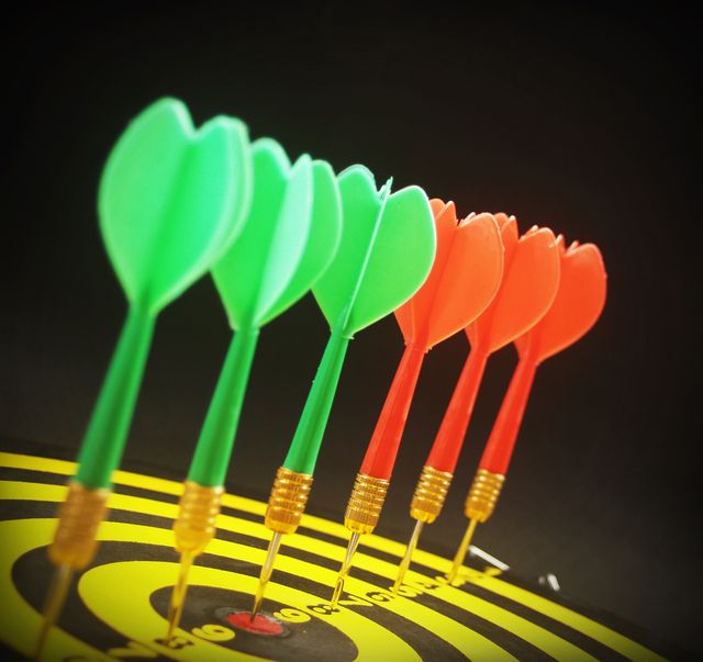 Six colorful darts, three green and three red, are perfectly landing on the bullseye of a target. The vibrant colors contrast with the black background, making the darts stand out prominently. This image is ideal for use in sports-related materials, advertisements promoting accuracy and precision, or content related to hobbies and leisure activities. It can also be used in presentations emphasizing focus and success.