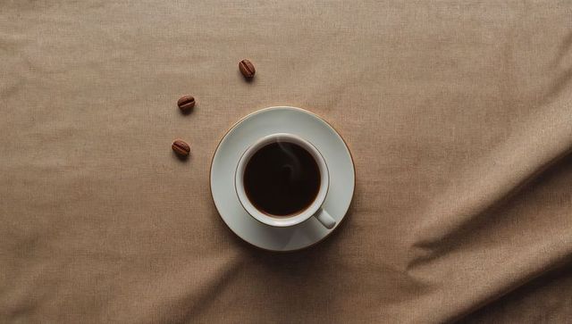 Steaming black coffee in white porcelain cup with gold rim on beige fabric flat lay