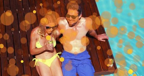 Sunbathing couple relaxing on wooden deck beside turquoise pool with cocktails and bokeh