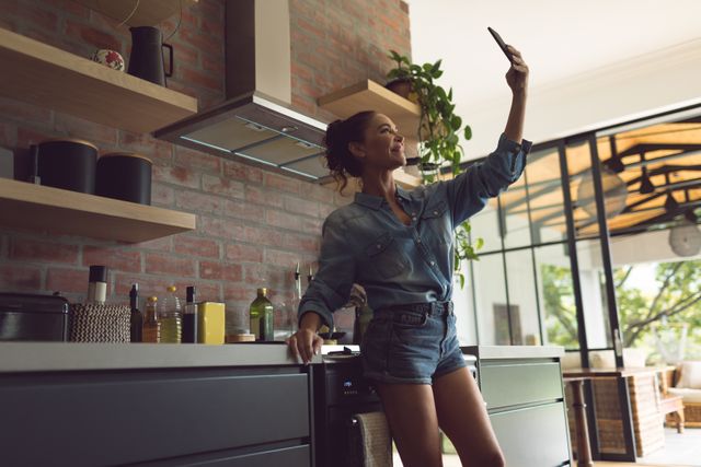 Woman taking selfie in modern kitchen, wearing casual denim outfit. Ideal for lifestyle, home decor, social media, and technology-related content.