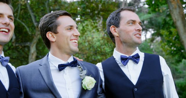 Three groomsmen smiling together in formal attire at an outdoor wedding. The men are dressed in white shirts, bow ties, and suits or vests, capturing a joyful moment. Ideal for use in wedding planning, event advertisements, friendship themes, or celebratory event materials. Perfect for illustrating concepts of companionship, joy, and wedding festivities.