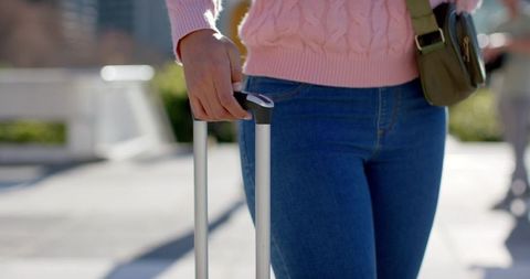 Woman walking with suitcase handle in sunny urban plaza wearing pink sweater and jeans