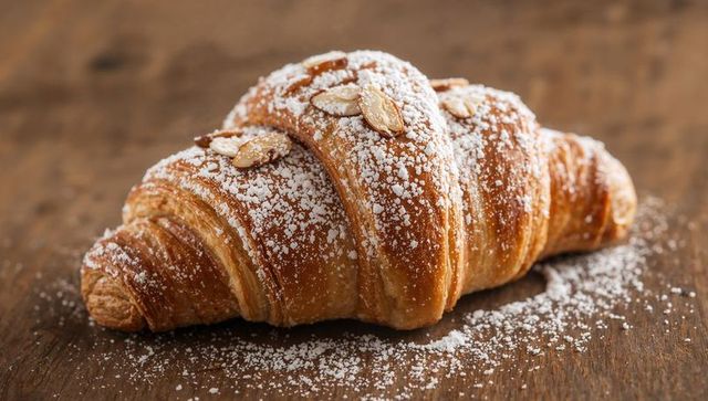 Buttery almond croissant sitting on rustic wooden table being dusted with powdered sugar