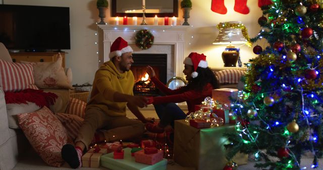 Couple with Santa hats exchanging gifts by Christmas tree and fireplace - Download Free Stock Images Pikwizard.com