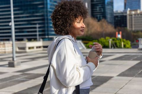 Urban woman holding reusable cup walking through modern plaza in sunny city morning