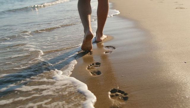Walking barefoot along golden shoreline leaving footprints in wet sand with gentle sea foam