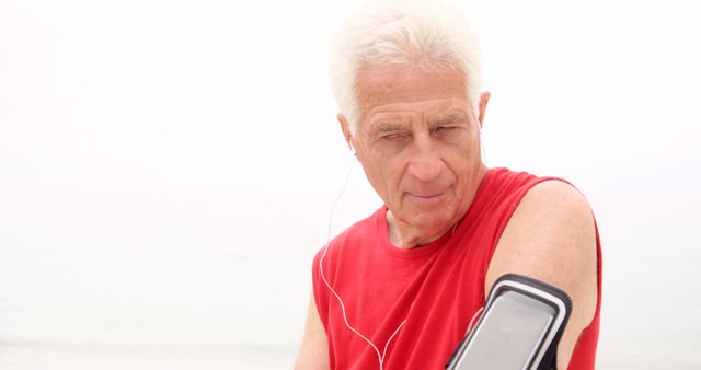 Elderly man outdoors wearing a red sleeveless shirt while using a smartphone arm band and listening to music or an audio guide through headphones. Ideal for themes centered on senior health, outdoor fitness, active aging, and workout routines for the elderly.