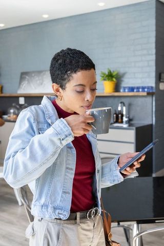 African american woman drinking coffee while checking smartphone in contemporary apartment kitchen