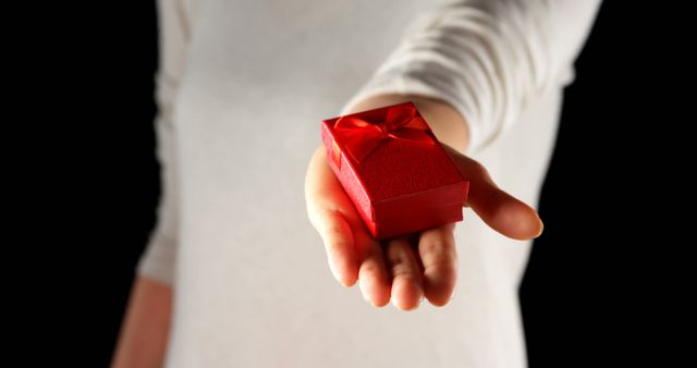 Close-up view of a hand offering a red gift box tied with a bow against a black background. This image can be used for themes related to gifting, surprises, celebrations, holidays, special occasions, and expressions of appreciation or love.