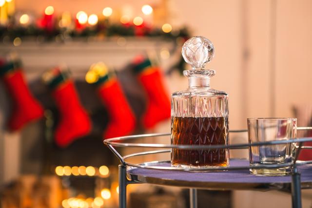 This image depicts a close-up of a whiskey bottle and glass on a table, with Christmas decorations in the background, including a fireplace with stockings. Ideal for use in holiday-themed advertisements, festive greeting cards, or articles about holiday celebrations and cozy home settings.