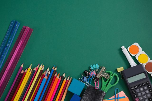 This image features a variety of school supplies arranged on a green background. Items include colored pencils, rulers, a calculator, scissors, paper clips, binder clips, and watercolor paints. Ideal for use in educational materials, back-to-school promotions, office supply advertisements, and creative projects.