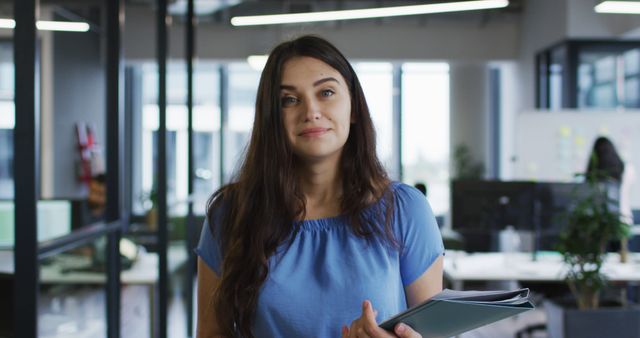 Confident businesswoman holding documents in a modern glass-walled office environment. Ideal for use in websites, articles, or materials related to business, professional settings, female leadership, corporate environments, career opportunities, and working professionals.