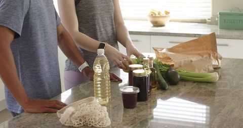 Diverse couple unpacking healthy groceries at sunlit kitchen island, sorting produce