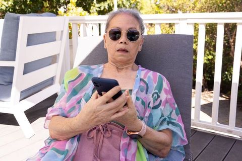 Senior asian woman relaxing on sun deck using smartphone, wearing sunglasses and colorful coverup