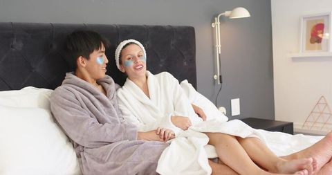 Diverse couple reclining on bed wearing plush robes and under-eye patches enjoying spa night