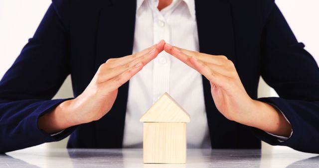A Caucasian woman in a business suit forms a protective gesture over a wooden house model, with copy space. Her hands symbolize security and care in the context of real estate or insurance services.