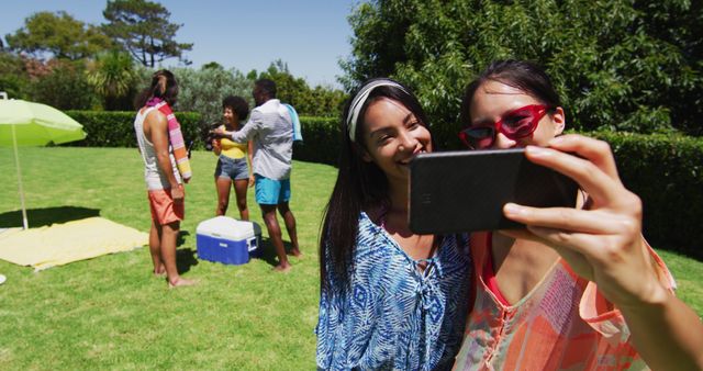 Group of friends enjoying fun outdoor summer party. Two people in foreground taking a selfie while others socialize in background. Scene captures youthful energy and casual setting, perfect for marketing summer events, friendship themes, and leisure activities.