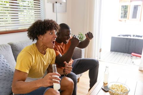 Young diverse friends cheering on sofa watching game holding drinks and popcorn