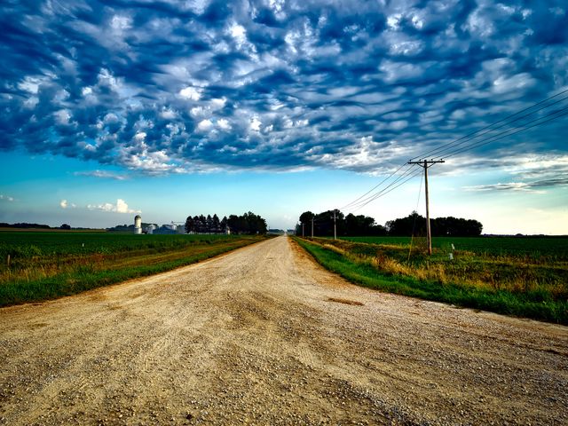 Gravel rural road stretching into distance, bordered by green fields and power poles. Dramatic clouds add character to the sky, indicative of impending weather. Ideal for use in travel, agriculture, and scenic landscape materials.