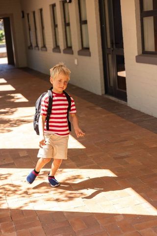 Young boy walking to school carrying backpack on sunlit brick walkway with long shadows